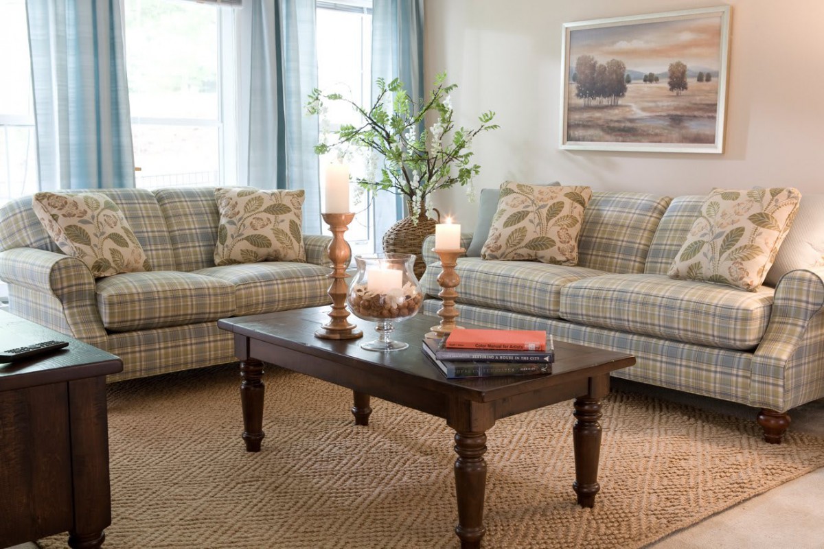 A cozy living room with plaid sofas, a wooden coffee table adorned with candles and books, complemented by a large window and wall artwork.