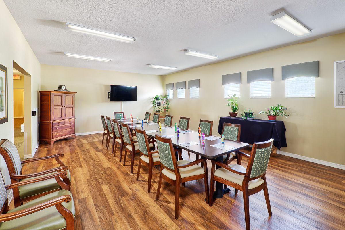 A well-lit conference room with a long table and multiple chairs, featuring a television and plants along the windows.