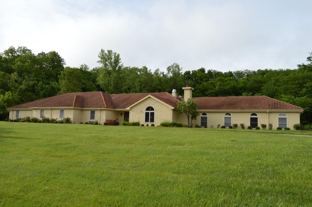 A single-story house with a red-tiled roof, surrounded by a large green lawn and trees in the background.