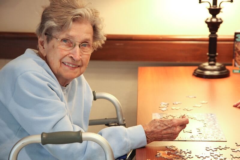 An elderly woman smiling while working on a puzzle indoors.