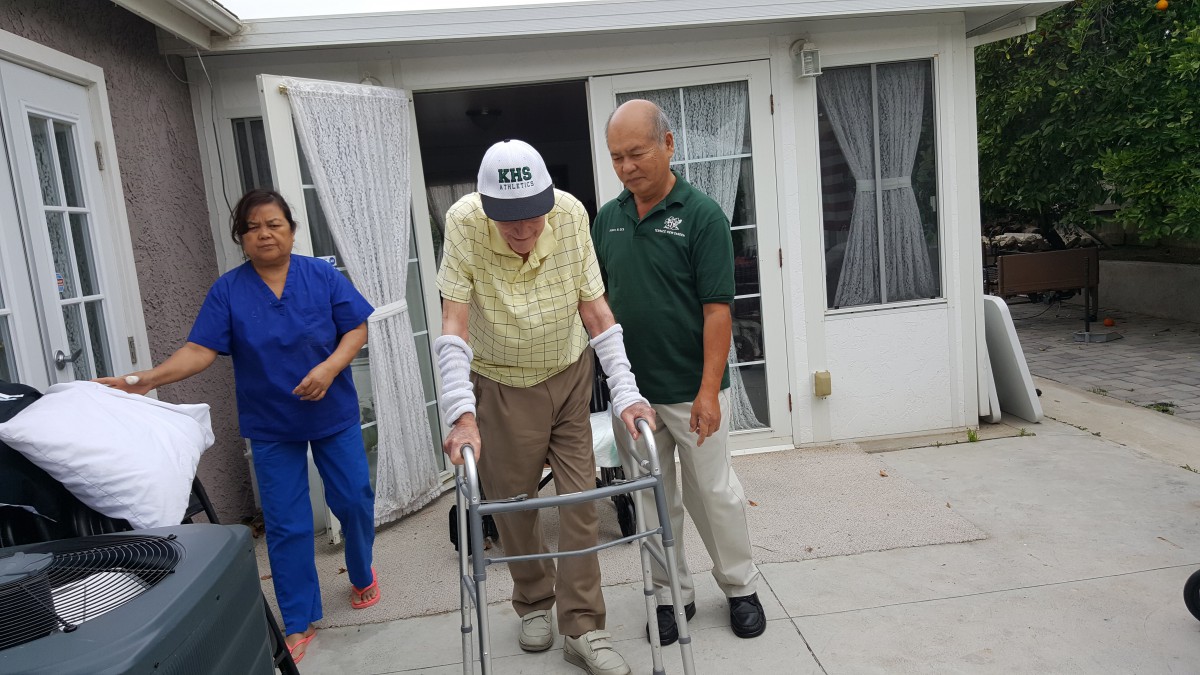 An elderly man using a walker is assisted by two caregivers outside a building.
