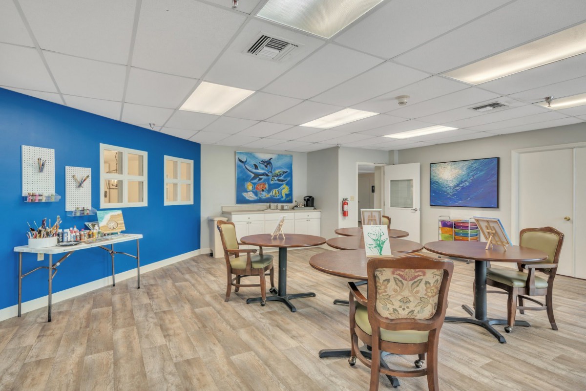 A brightly lit room with tables and chairs, featuring artwork on the walls and a wooden floor.