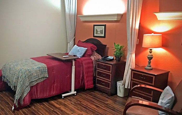 A cozy and well-furnished single hospital room with a wooden floor, bedside table, lamp, and curtains.