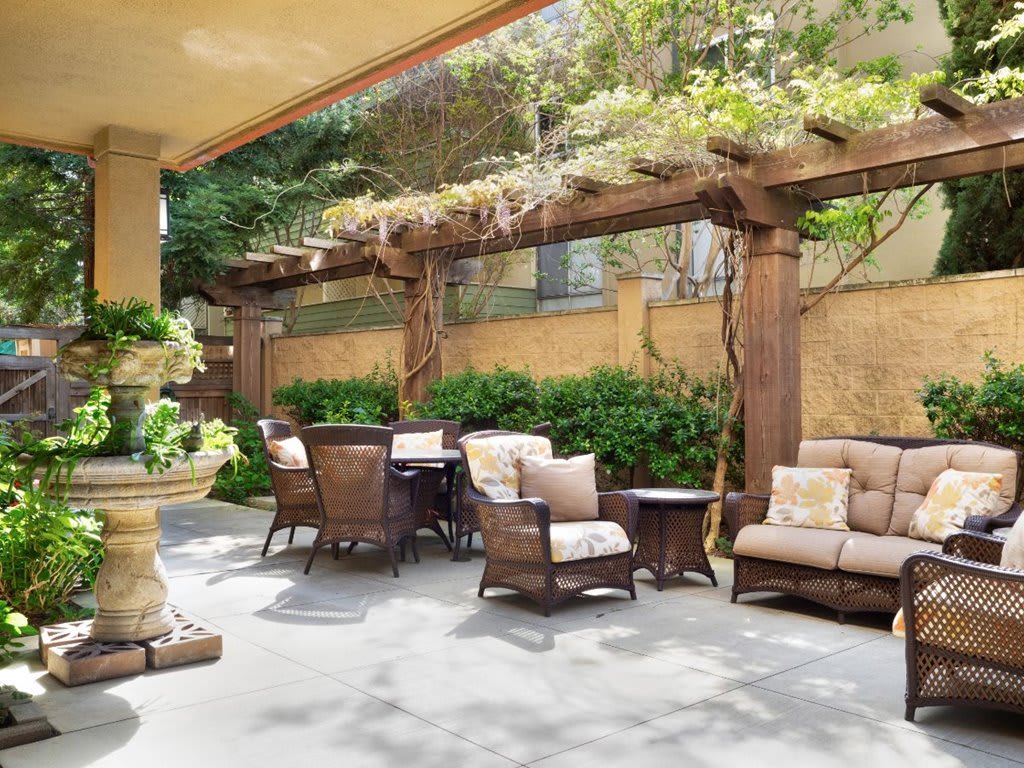 A cozy outdoor patio featuring comfortable seating under a wooden pergola, surrounded by lush greenery.