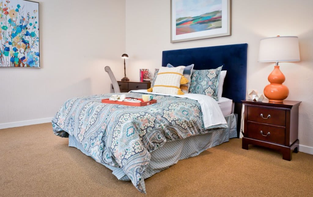A cozy bedroom featuring a bed with patterned bedding, a blue headboard, and a tidy nightstand with a lamp.