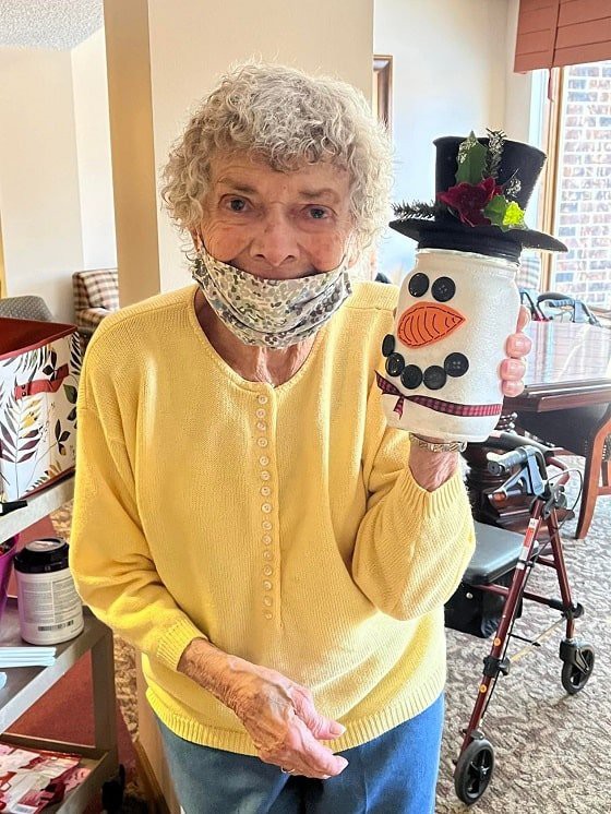 An elderly woman in a yellow sweater holds up a snowman craft indoors, with a cheerful expression.