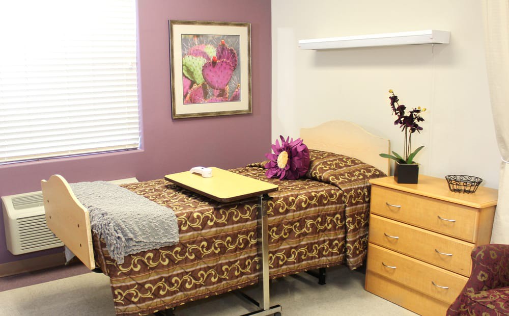 A hospital room featuring a patterned bedspread, a bedside table with flowers, and a vibrant painting on the wall.