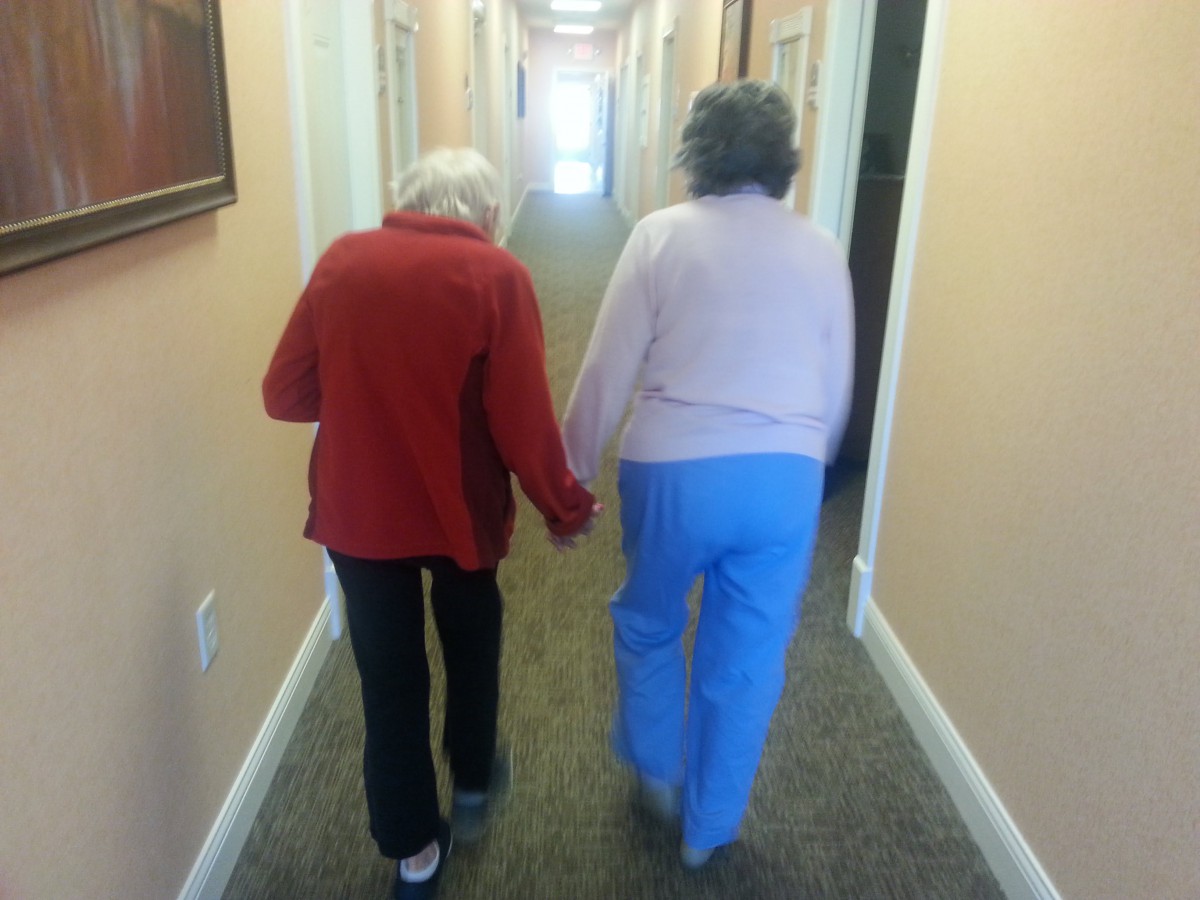 Two elderly individuals walking hand in hand down a hallway, symbolizing companionship and support.