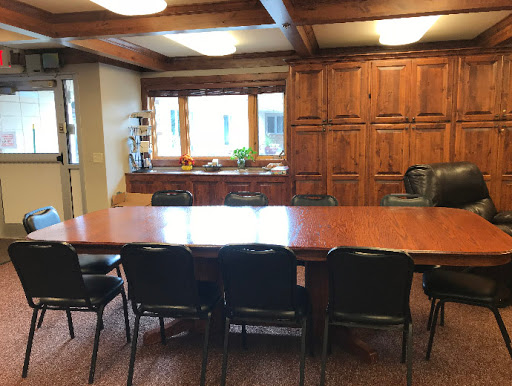 A spacious conference room featuring a large wooden table surrounded by black chairs, with natural light streaming in from the windows.