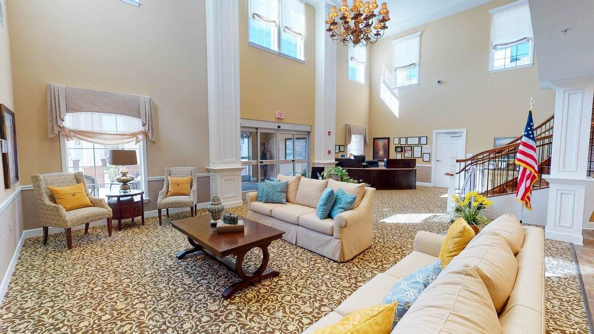 A spacious and elegantly decorated lobby with comfortable sofas, a central table, a large chandelier, and tall windows allowing natural light to fill the room.