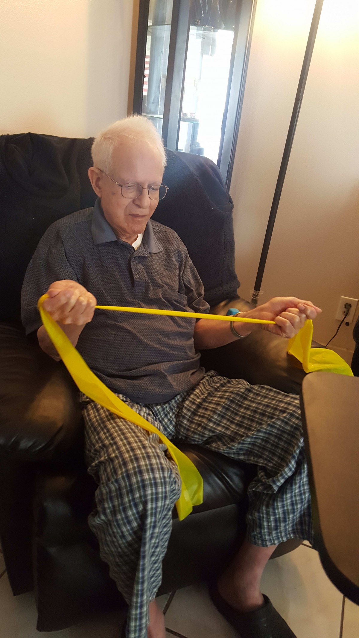 An elderly person seated indoors using a yellow resistance band for exercise.