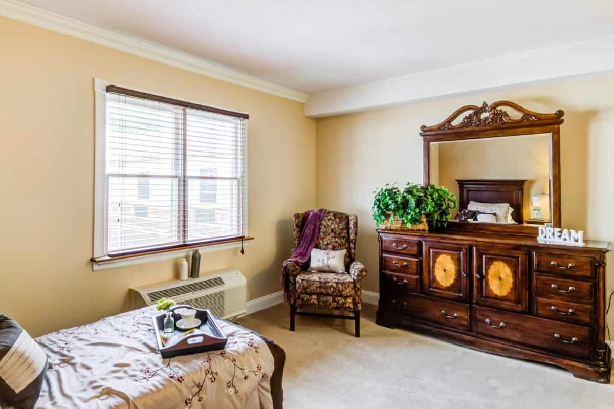 A cozy bedroom with elegant wooden furniture, featuring a bed, armchair, and dresser near a window.