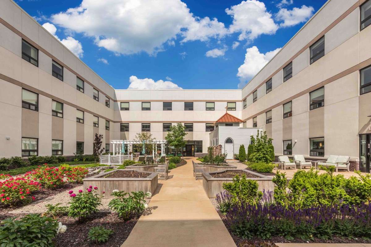 A well-maintained courtyard garden surrounded by a multi-story building under a blue sky with scattered clouds.
