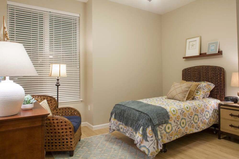 A cozy bedroom with a single bed, wicker chair, and a standing lamp beside a window with blinds.