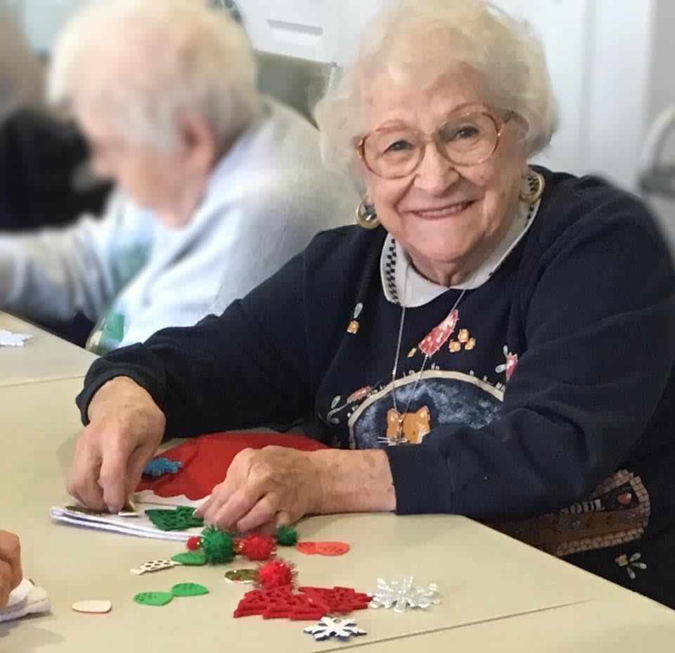 An elderly woman smiles while engaging in a crafting activity at a table filled with colorful materials.