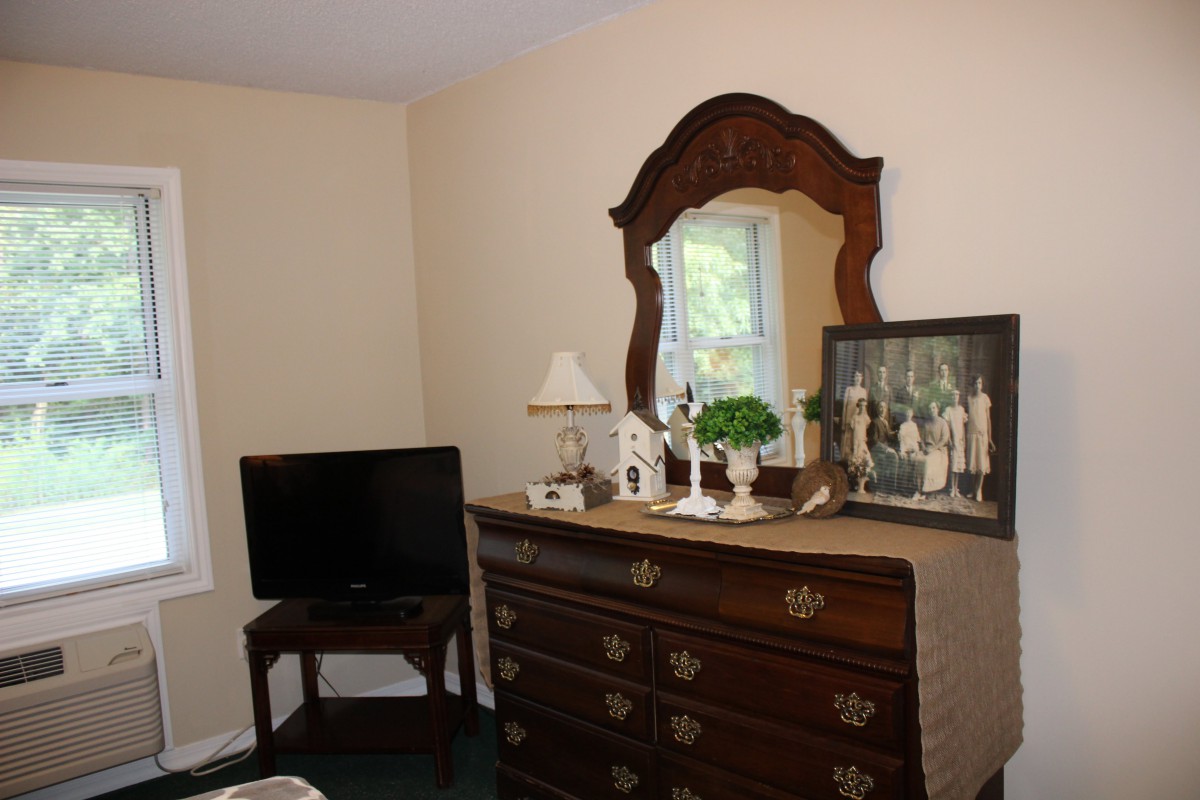 A cozy bedroom corner featuring a traditional wooden dresser with a mirror, decorated with lamps and plants, alongside a TV on a stand.
