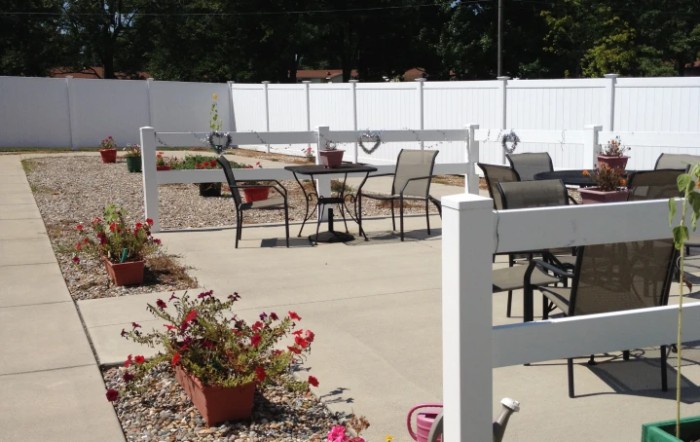 A sunny outdoor patio area with tables and chairs, surrounded by a white fence and decorated with potted flowers.