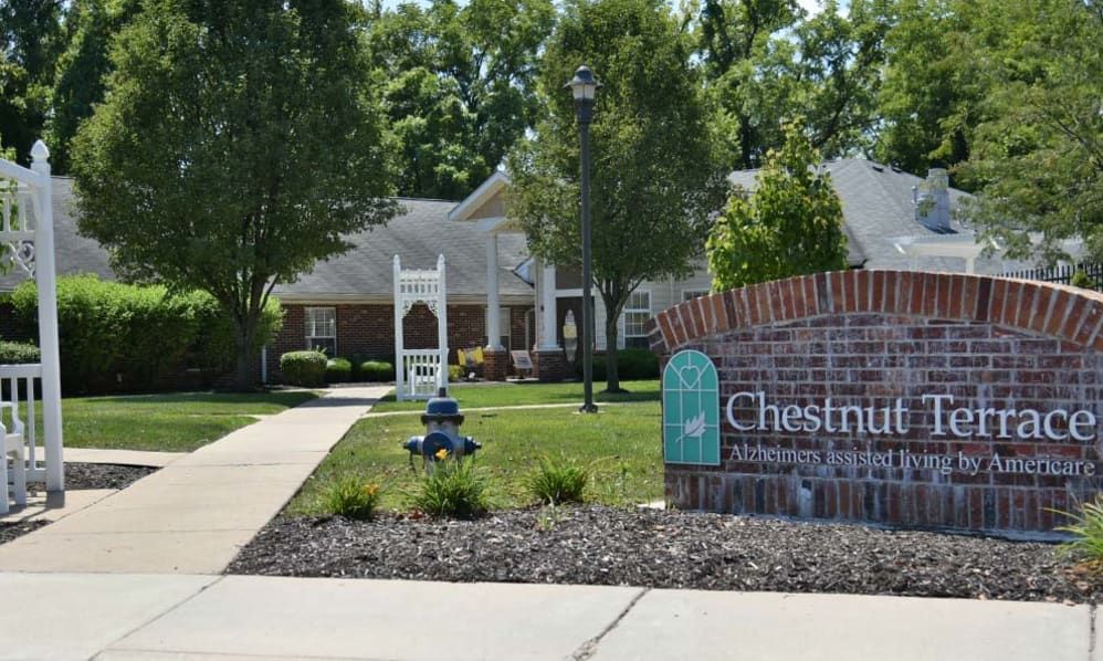 A residential complex with a brick sign in the foreground and a well-maintained landscape featuring trees and pathways.