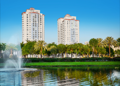 A scenic view of modern buildings beside a tranquil park with palm trees and a reflecting pond.
