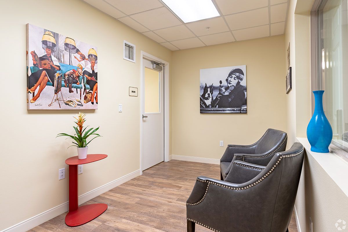 A cozy waiting room with two grey chairs, colorful artwork on the walls, and a small plant on a red table.