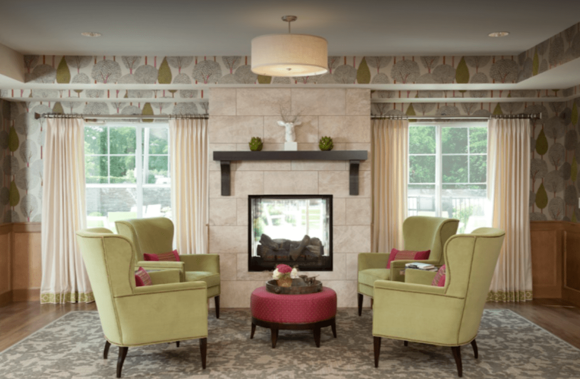 A cozy living room with green armchairs surrounding a central fireplace, complemented by a red ottoman and large windows.