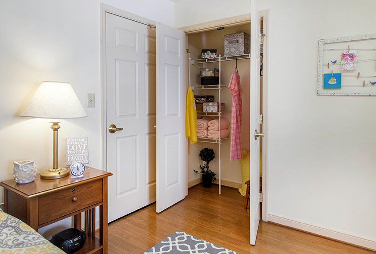 A cozy bedroom with an open closet showing clothes hanging neatly, a bedside table with a lamp, and a patterned rug on the wooden floor.