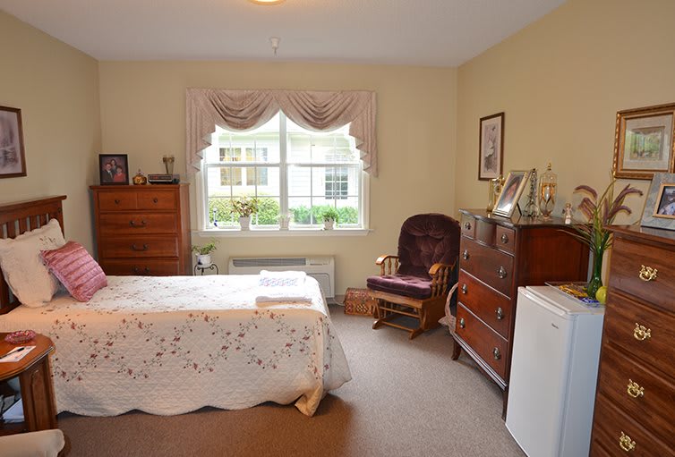 A cozy bedroom featuring a bed with a lace bedspread, a wooden dresser and chest of drawers, a rocking chair, and a window with draped curtains.