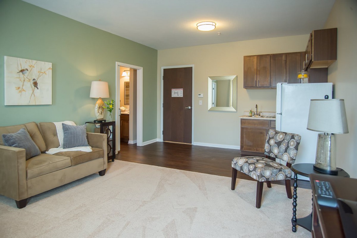 A cozy apartment interior featuring a living room with a sofa and chair, a kitchenette, and a softly lit lamp near the doorway.