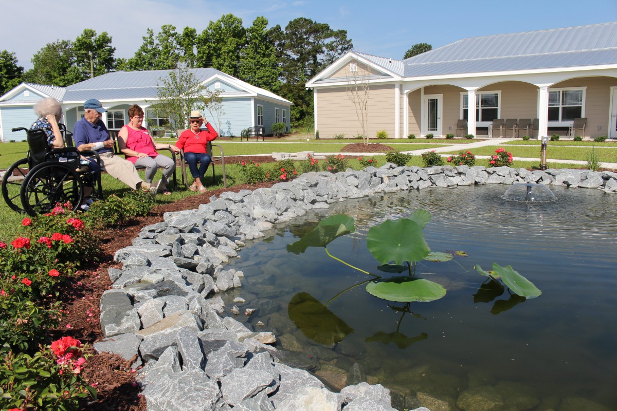 Two people sitting by a pond in a landscaped garden with houses in the background.