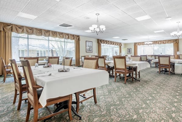 An elegant dining room with neatly arranged tables and chairs, decorated with white tablecloths and natural lighting from large windows.