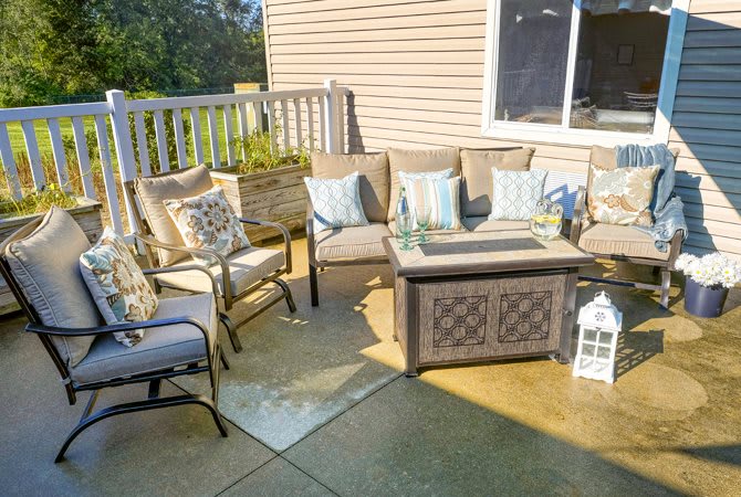 A cozy patio setup with outdoor furniture, cushions, and a fire pit table on a sunny day.