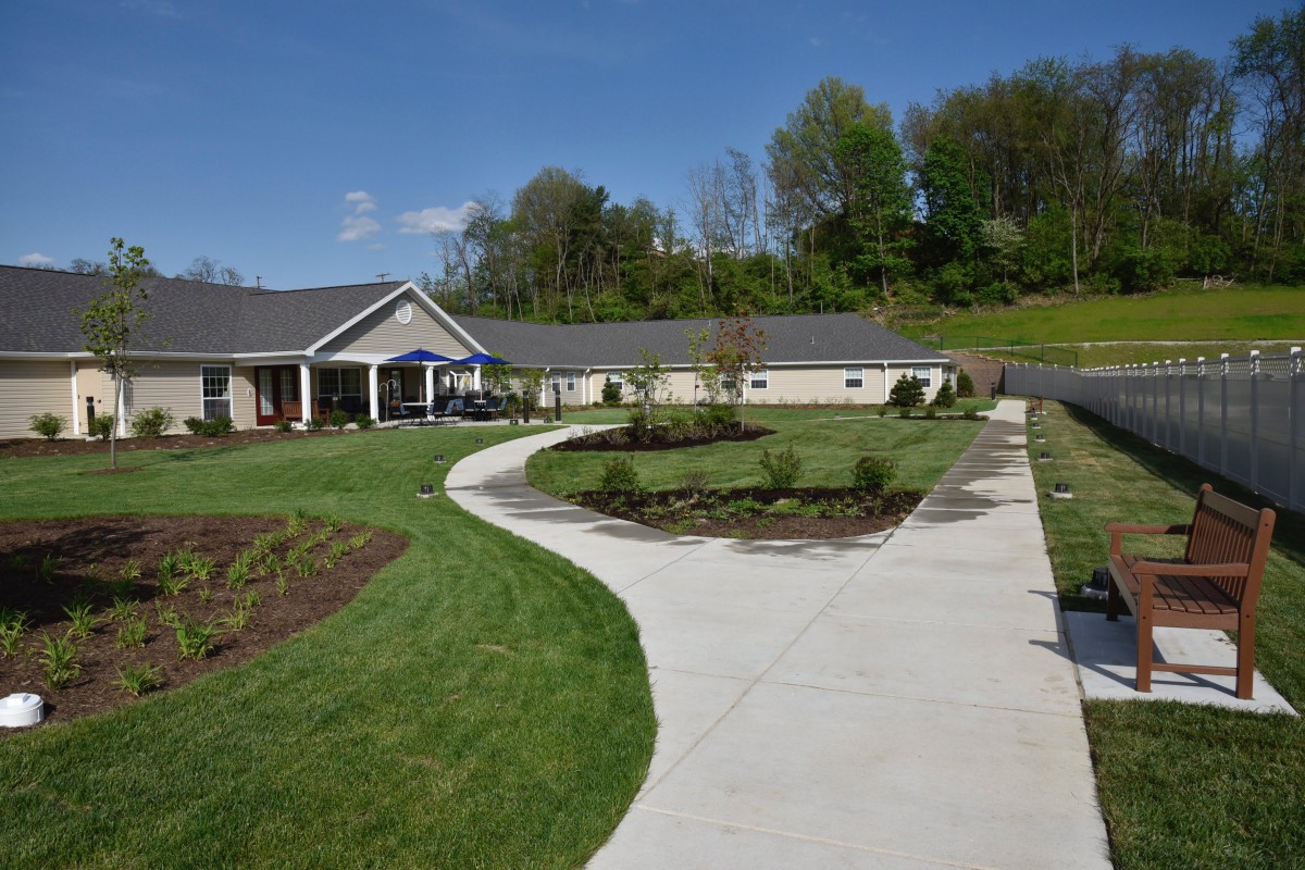 A single-story house with a curved pathway leading through a landscaped garden area with trees in the background under a clear blue sky.