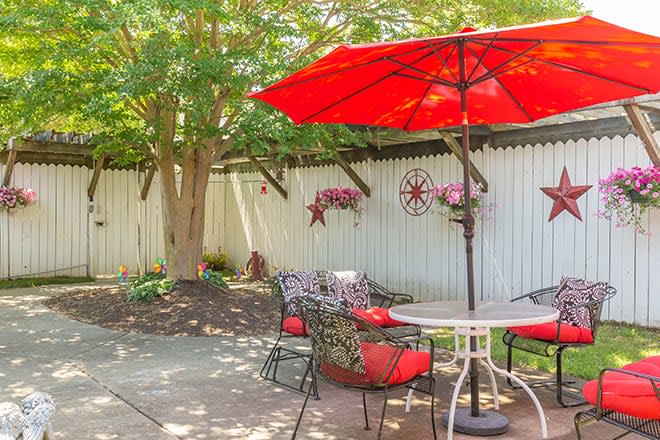 A cozy patio area with red-cushioned chairs under a red umbrella, surrounded by a landscaped garden and white fence.