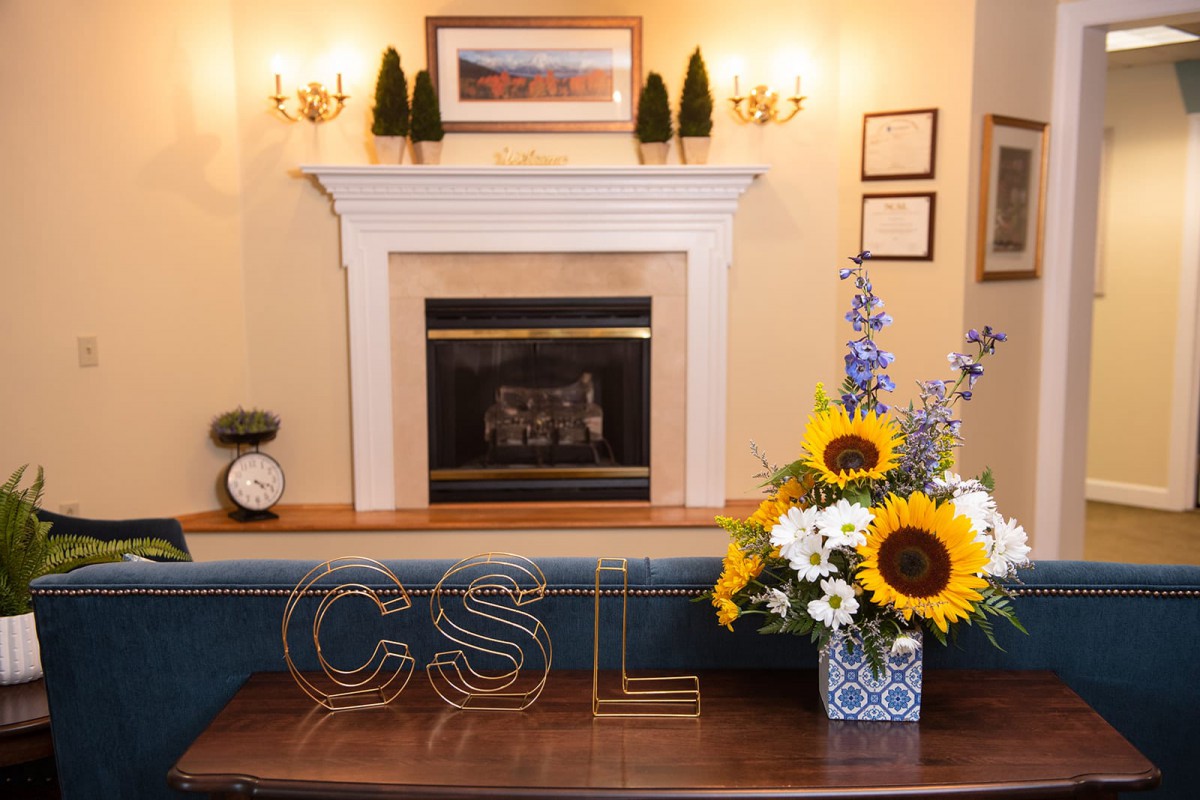 A cozy living room setup featuring a fireplace with framed artwork above, flanked by sconces and decorative pieces. A table in the foreground displays a vase with bright sunflowers and other colorful flowers.