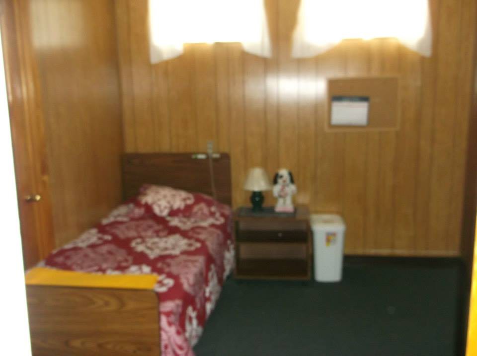 A small bedroom with wood-paneled walls, featuring a single bed with floral bedding, a side table, and a lamp.
