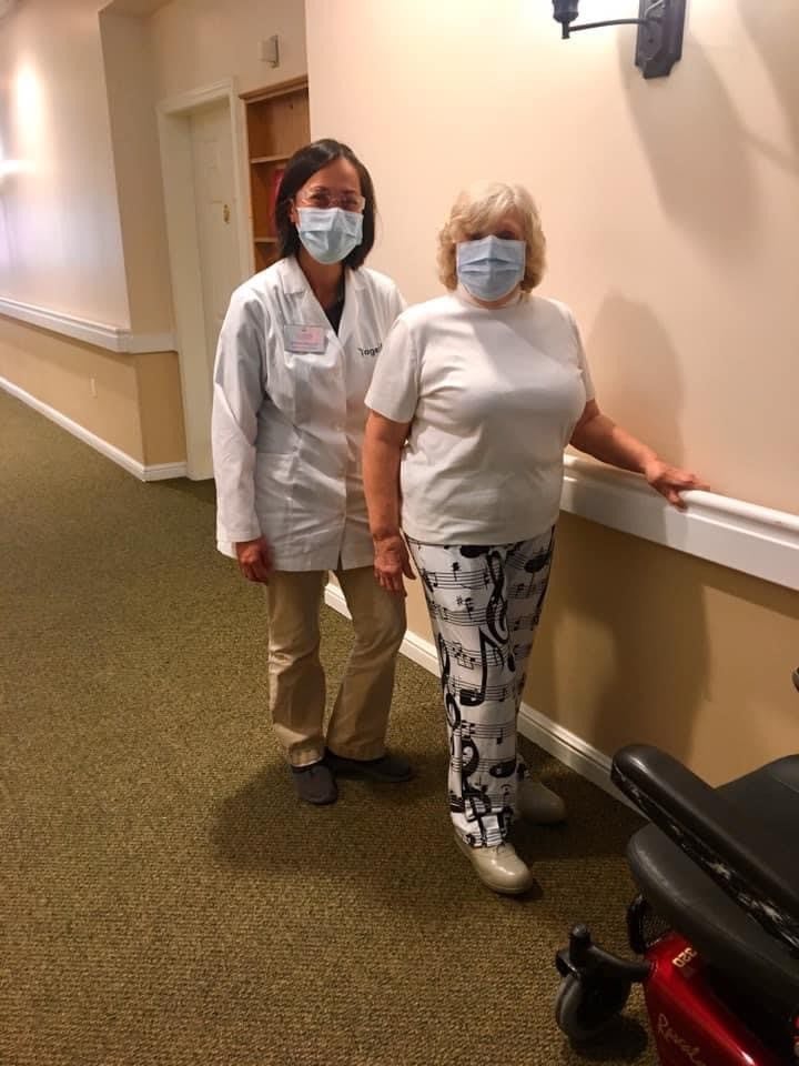 A healthcare professional assists a patient during rehabilitation in a facility, both wearing masks for protection.