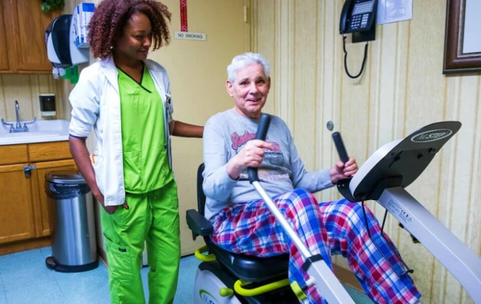 An elderly person using exercise equipment with the assistance of a healthcare worker.