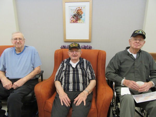 Three elderly men, possibly veterans, sitting together in a living room, wearing military hats.