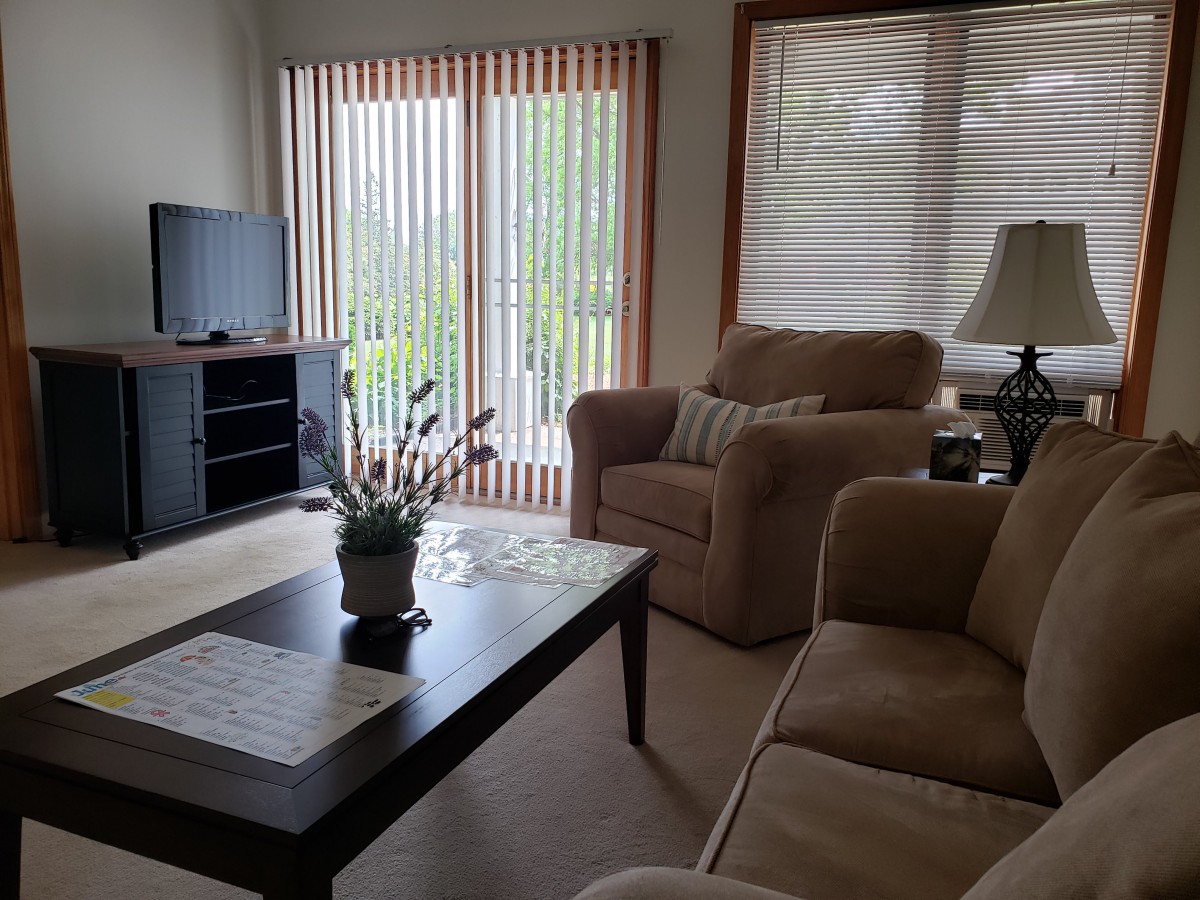 A cozy living room with a sofa set, coffee table, and television, featuring large windows with blinds.