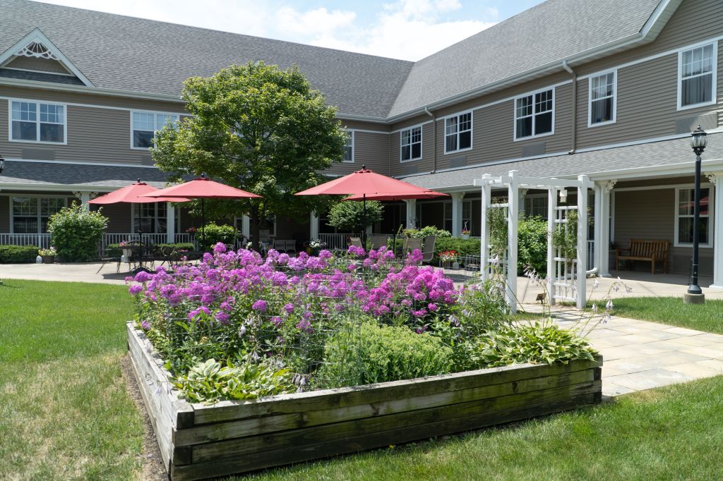 A beautiful garden with vibrant flowers and a patio area featuring red umbrellas beside a large building.