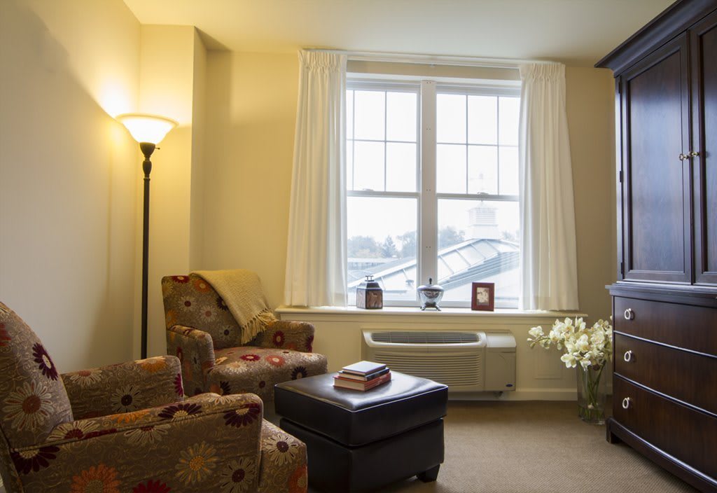 A cozy living room interior with an armchair, ottoman, and a large window letting in natural light.