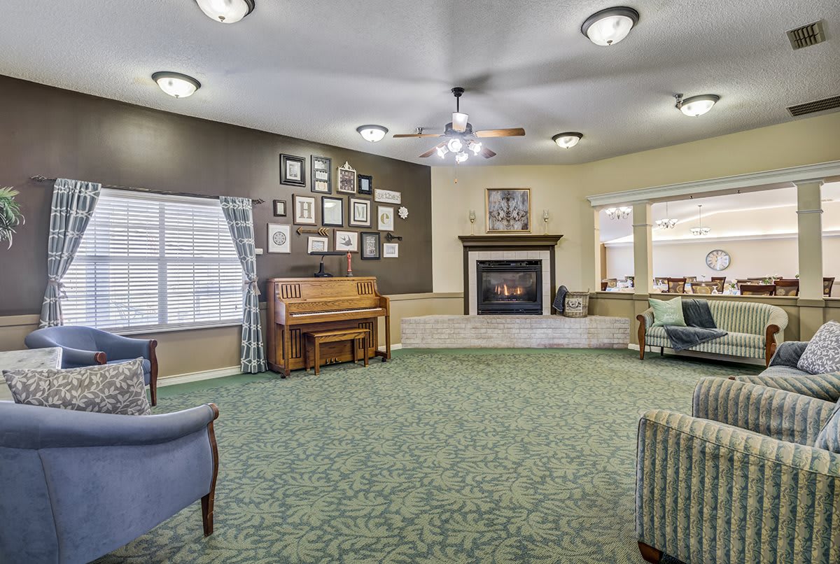 A cozy living room featuring a piano, fireplace, and seating area with green carpet and framed pictures on the wall.