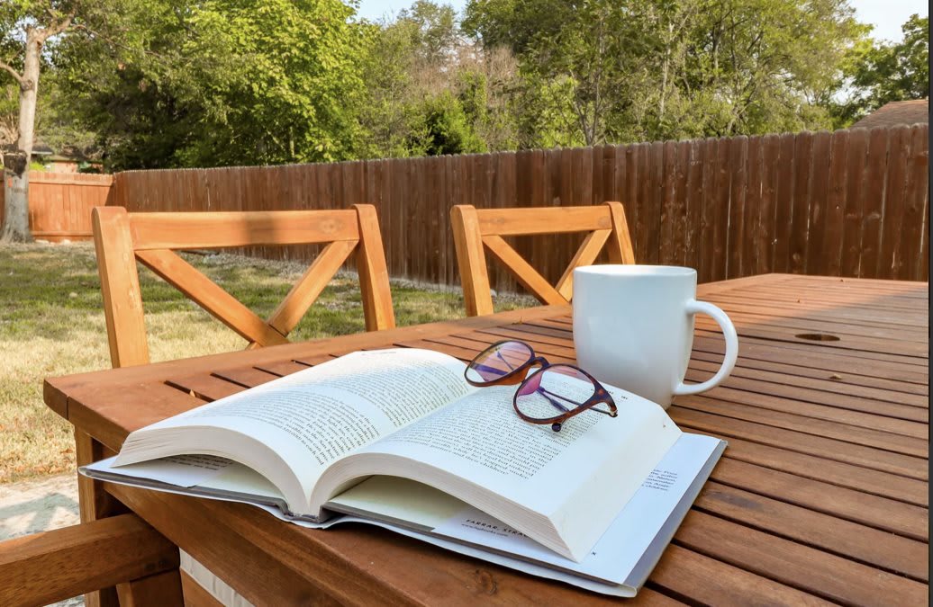 A relaxing outdoor reading setup with an open book, glasses, and a coffee cup on a wooden table.