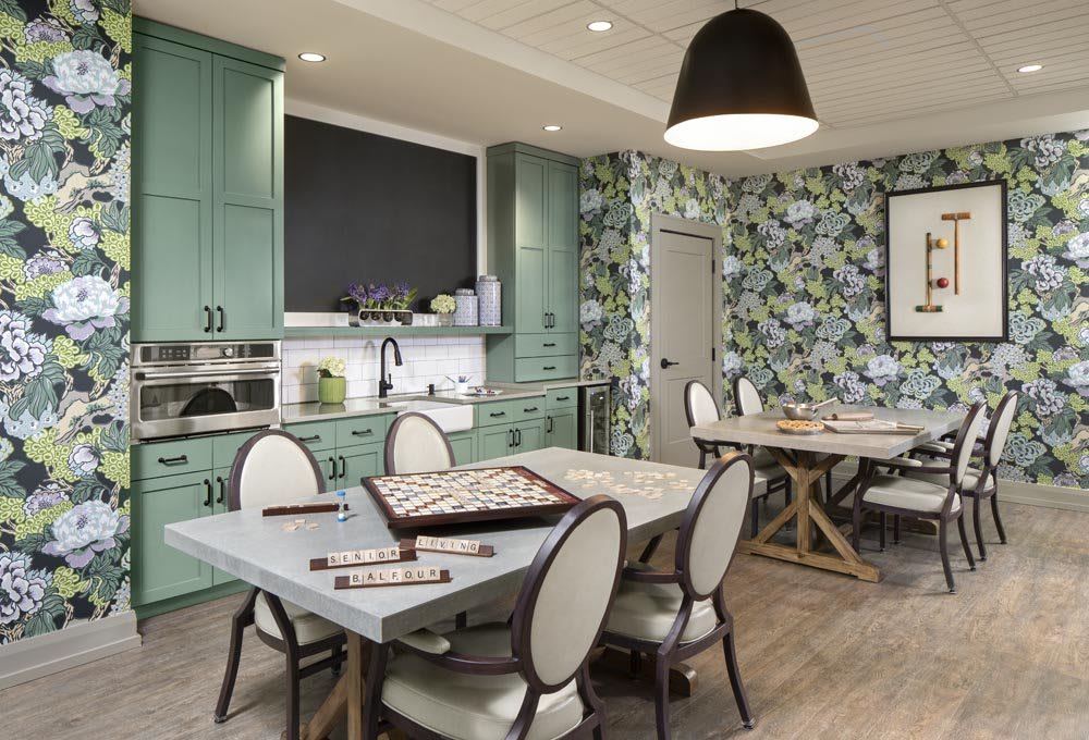 A modern kitchen with green cabinets and floral wallpaper, featuring a dining table set up for a board game.