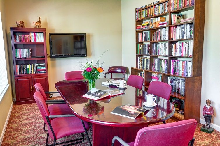 A conference room with a polished wooden meeting table surrounded by red chairs, bookshelves filled with books, a TV mounted on the wall, and a vase of flowers on the table.