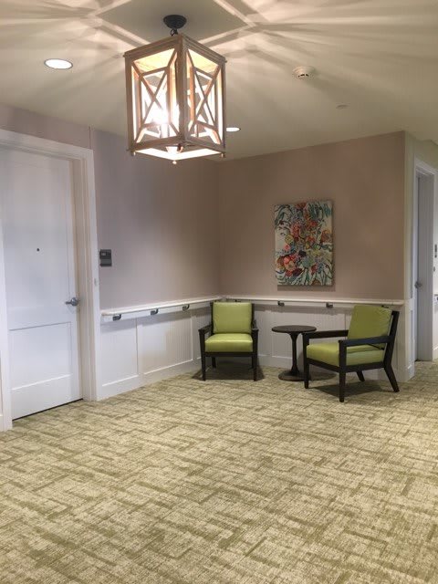 A cozy seating area with two green chairs and a small table set against a beige wall, adorned with artwork and illuminated by an overhead lamp.