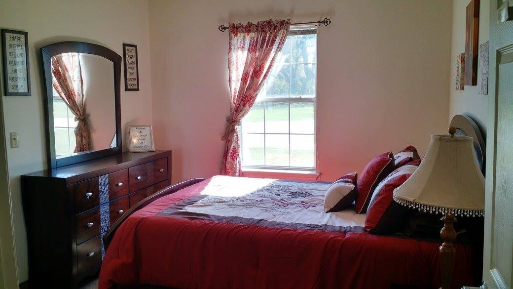 A cozy bedroom with a red bedspread, wooden dresser, and sunlight streaming through the window.