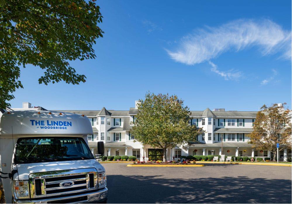 A large white building with multiple windows and a tree in front, next to a parked vehicle under a clear blue sky with wispy clouds.