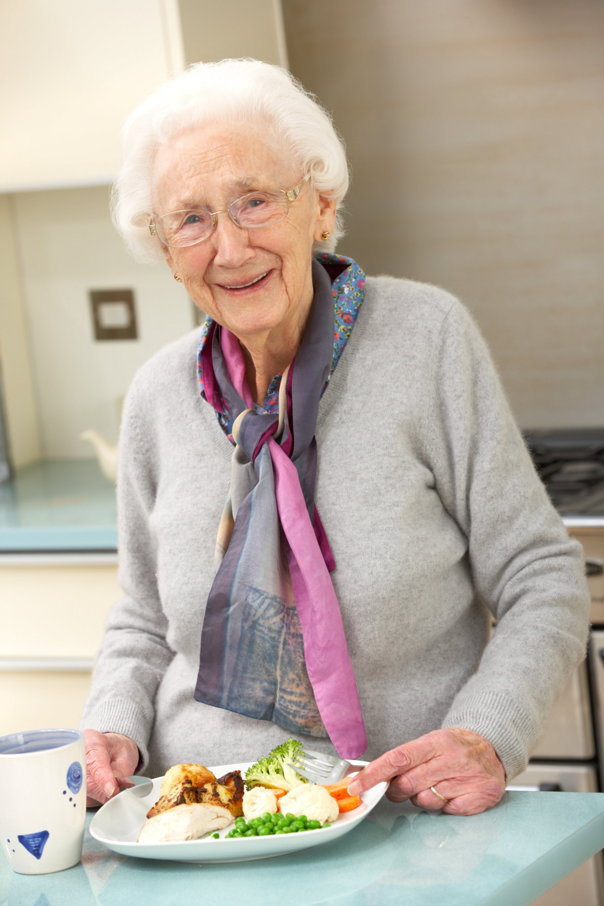 An elderly woman smiles warmly while standing in a kitchen, wearing a grey outfit and a colorful scarf.