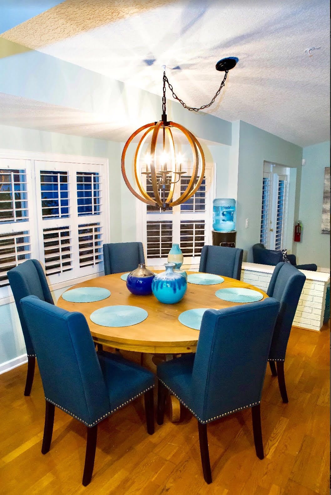 A cozy dining room with a round wooden table, surrounded by blue upholstered chairs and illuminated by a modern chandelier.